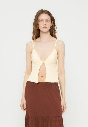 Jeune femme aux longs cheveux portant un haut spaghetti crème avec fente frontale et une jupe marron taille haute, debout devant un fond uni.