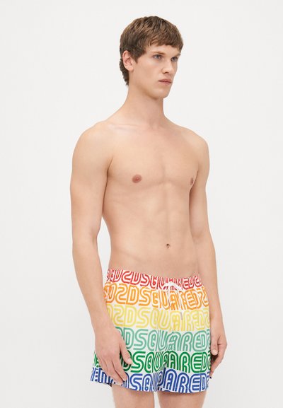 En shirtless ung man som bär färgglada shorts med upprepande "DSQUARED2"-text i röd, orange, gul, grön och blå, stående mot en vit bakgrund.