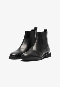 Botas Chelsea de piel negra con paneles laterales elásticos, punta redonda, lengüetas de tiro y suela plana de goma. Textura suave con costuras mínimas.