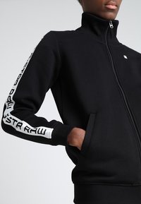 Persona que lleva una chaqueta negra con cremallera y texto de marca en blanco a lo largo de las mangas, con una mano en el bolsillo, recortada en el cuello y la cintura.