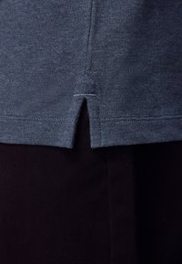 Modrý texturovaný lem košile s bočním rozparkem přes černé kalhoty zobrazený zblízka.