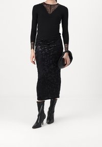 Haut noir à manches longues avec encolure en dentelle, associé à une jupe texturée noire à motifs floraux et des bottes noires pointues montant jusqu'aux genoux, tenant une pochette noire.