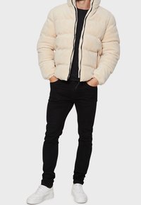 Chaqueta de plumas de color beige claro y suave, con un cuello alto y cremallera negra. Llevada sobre una camiseta negra con pantalones ajustados negros y zapatillas blancas.