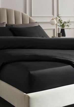 Bett mit schwarzer Bettwäsche, beigem gepolstertem Kopfteil, Nachttisch mit moderner Messinglampe und Vase mit grünen Zweigen, weiße vertäfelte Wand im Hintergrund.