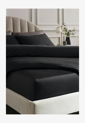 Bett mit schwarzer Bettwäsche, beigem gepolstertem Kopfteil, Nachttisch mit moderner Messinglampe und Vase mit grünen Zweigen, weiße vertäfelte Wand im Hintergrund.