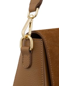 Hoek van een bruine leren tas met een suède textuur. Bevat een gouden metalen sluiting en een tan kleurige strap bevestiging. Schone stikseldetails.