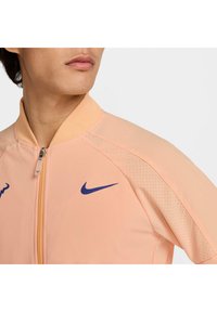 Chaqueta Nike en durazno claro, con cierre delantero, cuello acanalado y logo morado. Los detalles incluyen tela perforada en los hombros.