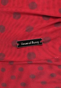 Crvena mrežasta tkanina s tamnim točkicama. Sadrži pravokotni srebrni oznaku s ugraviranim "House of Sunny". Mekana tekstura i lagani dizajn.