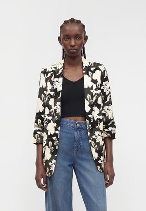 Femme aux cheveux tressés portant un top court noir, un blazer floral noir et blanc, et un jean bleu taille haute, debout devant un fond uni.