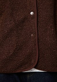 Brun fleecetröja med texturerad yta, tryckknappar och kontrasterande kantband längs kanterna, lämplig för vardagsbruk.