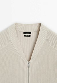 Pull en maille beige côtelée avec une fermeture éclair argentée à l'avant et un col en V, avec une étiquette de marque noire à l'intérieur de l'encolure.