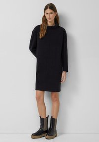 Czarna, oversize sukienka z wysokim kołnierzem i długimi rękawami. Gładka faktura i prosta sylwetka. W połączeniu z czarnymi botkami za kostkę.