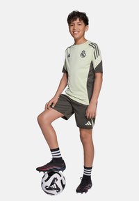 Lichtgroen en donkergrijs voetbalshirt met logo en zwarte accenten, gecombineerd met bijpassende shorts. Zwarte en rode voetbalschoenen, staand op een zwart-witte voetbal.