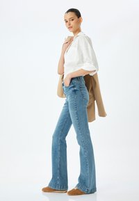 Vaqueros acampanados de un azul claro con cintura alta y desvanecidos, combinados con una camisa blanca de botones y un abrigo exterior beige. Un calzado marrón completa el look.