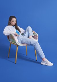 Sedia intrecciata colorata con una cornice gialla, modello che indossa una camicia bianca e jeans azzurri, accentuato da scarpe da ginnastica bianche con motivo su sfondo blu.