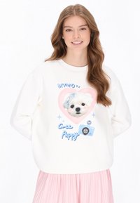 Weißes Sweatshirt mit einem Herzmotiv, das das Gesicht eines Welpen zeigt, blaue Akzente und den Text „Süßer Welpe“. Getragen mit einem pinken plissierten Rock.