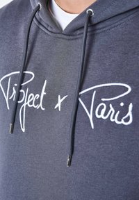 Sudadera gris con texto bordado en blanco "Project x Paris." Cuenta con cordón ajustable y aglets de metal en la capucha. Textura de tela suave.