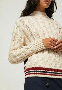 Strickpullover in Creme, mit geripptem Halsausschnitt und Bündchen, sowie einem gestreiften Saum in Rot, Marine und Creme. Strukturiertes Muster über das gesamte Stück.