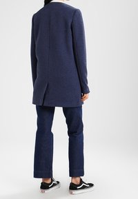 Marinblå texturerad lång kappa med en slits bak, kombinerad med mörkblå raka jeans och svarta sneakers med vita detaljer.