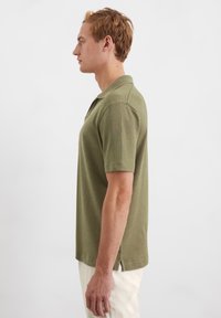 Olivgrünes Poloshirt mit klassischem Kragen, kurzen Ärmeln und Seitenschlitzen. Hergestellt aus weichem, strukturiertem Stoff. Getragen mit hellen Hosen.