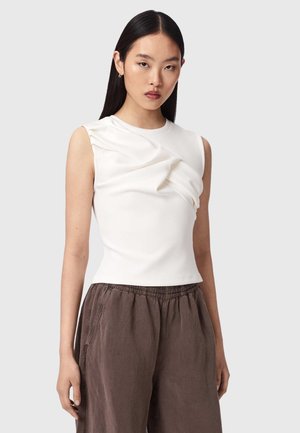 Jonge vrouw met lang steil haar, een witte mouwloze top met geplooide details en een losse bruine broek, staand tegen een grijze achtergrond.