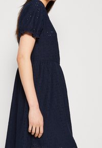 Vestido azul marino con ojales, mangas abullonadas, tejido texturizado, falda acampanada y sutiles patrones de recorte. Mostrado de lado.