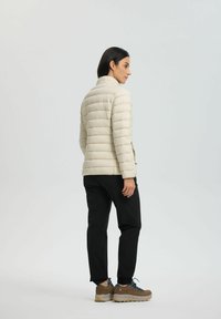 Veste matelassée beige clair avec un motif de piqûre horizontal, col haut, associée à un pantalon noir et des chaussures de randonnée marron.