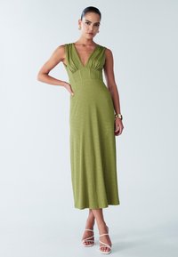 Groene jurk tot op de knie met een diepe V-hals, geplooide taille en een aangesloten taille. Gestructureerde stof en minimalistische witte sandalen met bandjes.