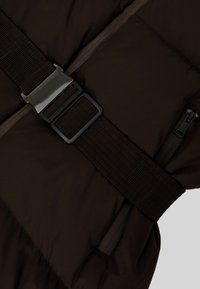 Gilet imbottito marrone con una larga fascia nera testurizzata e fibbia metallica, con tasche con cerniera inclinate su ciascun lato.