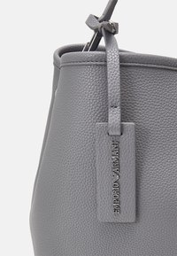 Harmaa nahkainen ämpärilaukku, jossa on teksturoitu pinta, varustettu yläkahvalla ja suorakulmaisella tarralla, johon on painettu "Emporio Armani."
