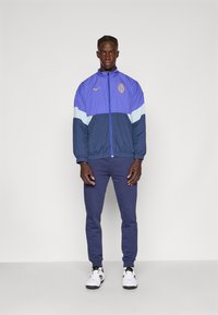 Violeta un tumši zilas vēja jaka ar rāvējslēdzēju priekšā, logo, elastīgām piedurknēm, kombinēta ar tumši zilām sporta biksēm un baltām sporta apaviem.