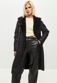 Sort frakke i imiteret læder med en blød, sort krave, enkeltspændt design og en lang silhuet, kombineret med sorte læderbukser.