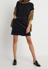Zwarte kortgebouwde top gecombineerd met een donkere corduroy rok, voorzien van een zak. Het model draagt zwarte instappers en een groene cardigan.