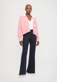 Rosa gerippter Cardigan mit braunen Knöpfen, weißem Kragenoberteil und marineblauen ausgestellten Hosen. Das Model trägt cremefarbene spitze Flats.
