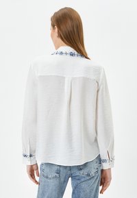 Camicia bianca con bottoni, caratterizzata da un tessuto testurizzato, dettagli blu ricamati sul colletto e sui polsini, e una vestibilità rilassata con orlo arrotondato.