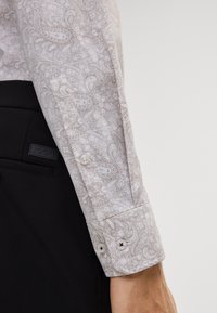 Langarmhemd in hellbeige mit weißem Paisleymuster, mit Knopfmanschetten und einem leichten Glanz. Schwarzer Stoff am unteren Ende sichtbar.