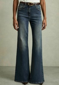 Jeans évasés bleu marine avec une taille moyenne, ceinture marron avec une boucle ovale, cinq poches et un léger dégradé sur les cuisses. La texture principale est le denim.