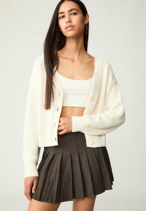 Cardigan ivoire en tricot avec une texture côtelée, doté de grands boutons, associé à une jupe brune plissée avec des fines rayures.