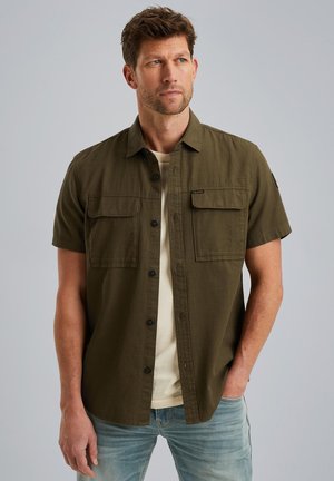 Camisa de botones de manga corta color verde oliva con dos bolsillos frontales, de tela ligera y un corte relajado, usada sobre una camiseta de color claro.