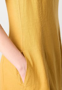 La main d'une personne repose dans la poche latérale d'une robe en lin jaune moutarde avec des détails de couture visibles.