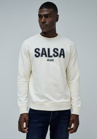 Sudadera crema con la inscripción "SALSA JEANS" en letras negras en negrita, cuello redondo y puños acanalados. Combinada con jeans de denim oscuros.