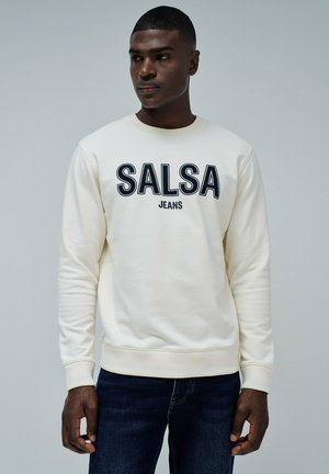 Sudadera crema con la inscripción "SALSA JEANS" en letras negras en negrita, cuello redondo y puños acanalados. Combinada con jeans de denim oscuros.