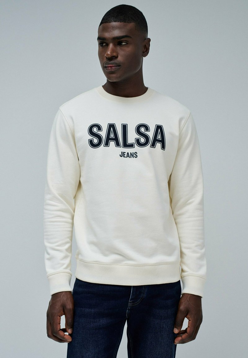 Sudadera crema con la inscripción "SALSA JEANS" en letras negras en negrita, cuello redondo y puños acanalados. Combinada con jeans de denim oscuros.