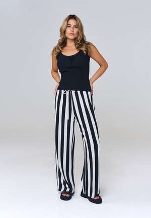 Mujer con el pelo largo y ondulado, vestida con una camiseta negra sin mangas y pantalones anchos de rayas verticales en blanco y negro, de pie con las manos en las caderas.