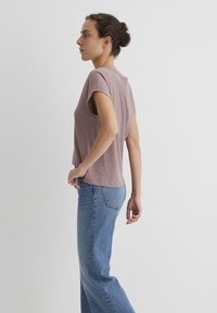 T-Shirt aus hellbraunem Stoff mit kurzen Ärmeln, kombiniert mit hoch taillierten blauen Jeans. Das Shirt hat eine lockere Passform und einen abgerundeten Saum.