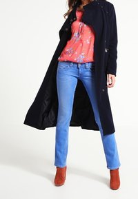 Námořnický trenčkot přes korálovou květinovou halenku, doplněný o světle modré džíny a oranžové kotníčkové boty. Hladké textury a přiléhavý střih.