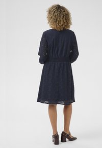 Abito di pizzo blu navy con maniche lunghe, vita stretta e gonna che arriva sopra il ginocchio. Indossato con scarpe a tacco quadrato marroni.