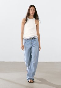 Hvid ærmeløs tanktop kombineret med lyseblå vide jeans og et mønstret bælte. Modellen bærer sorte sandaler. Ensfarvet baggrund.