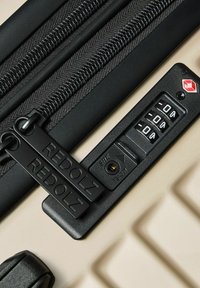 Schwarzer Reißverschluss und Schließmechanismus auf einem beige-farbenen Koffer. Das Schloss verfügt über drei Ziffern, die auf Null eingestellt sind, mit "REDOLZ" auf den Reißverschluss-Anhängern geprägt. Strukturierten Oberfläche.