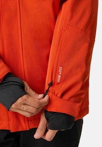 Giacca arancione brillante con dettagli neri, caratterizzata da un polsino con zip e il dettaglio in rilievo "STX™ 8000". Texture liscia, vestibilità sartoriale.